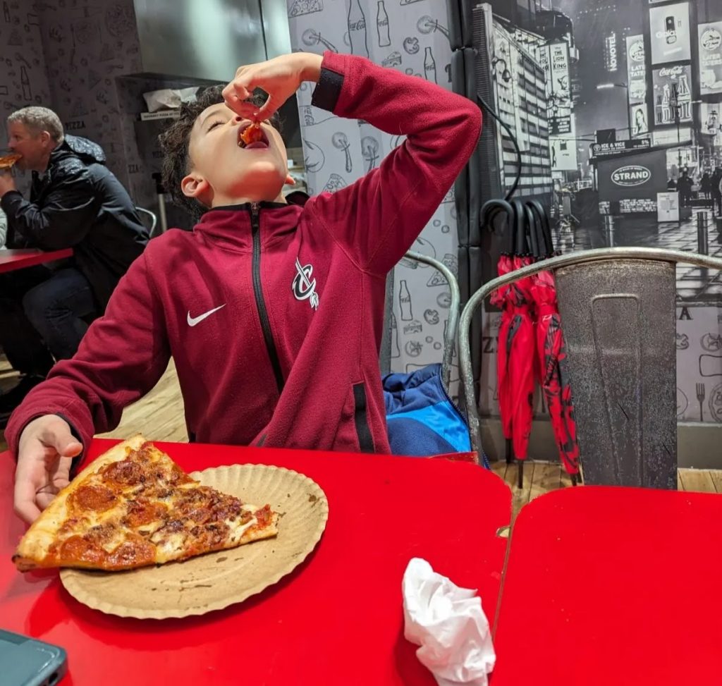Kid devouring New York style pizza from a brown paper plate. 